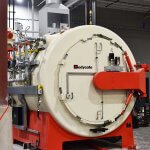 Large Bodycote vacuum furnace with round door, pipes and controls, on red base in a North American industrial facility.