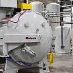 Bodycote vacuum furnace in a manufacturing plant, surrounded by pipes, tanks, control panels and a workstation with monitor.