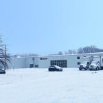 Bodycote industrial facility in snow, with company sign, parked cars and delivery lorry in front, leafless trees, clear sky.