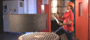 Worker in safety gear uses a hoist to lift a metal alloy part in an industrial facility; clipboard, remote, and red doors visible.