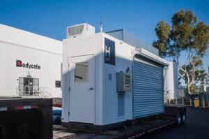 White hydrogen generator marked H gen on trailer by Bodycote thermal processing site, with large trees and clear sky in background.