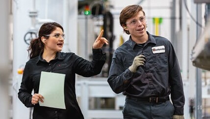 Two people in safety glasses and work uniforms walk through an industrial facility; one holds paper and gestures, the other listens.