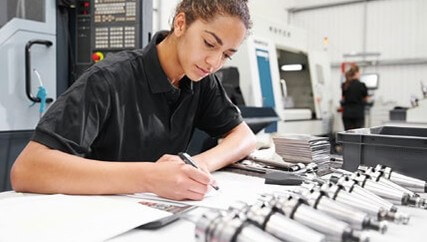 Person in a black polo shirt writing on paper at a workbench with metal parts in a Bodycote facility.
