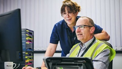 Two people in an office; one seated at a desk in a hi-vis vest viewing a computer monitor, another standing and looking at the screen.