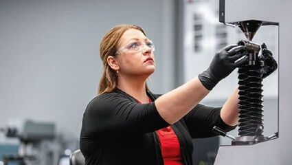 Woman in safety glasses and black gloves operates hardness testing machine, adjusting a metal part in a laboratory setting.