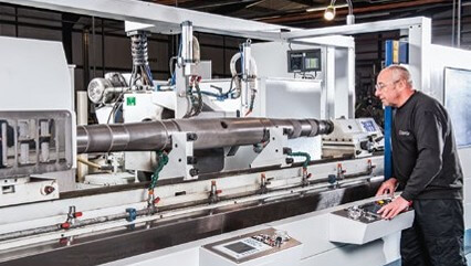 Man operating industrial grinding machine in a factory, monitoring controls as a long metal cylinder is processed; control panels visible.