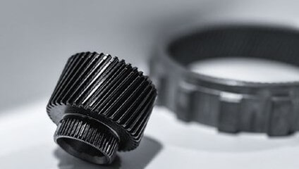 Two black metal gears on a white surface, one standing upright and one flat in the background, both with clearly visible teeth.