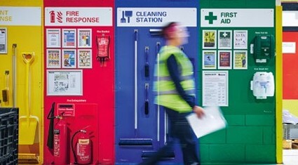 Worker in high-visibility vest walks past a wall with red, blue, and green zones for fire response, cleaning station, and first aid.