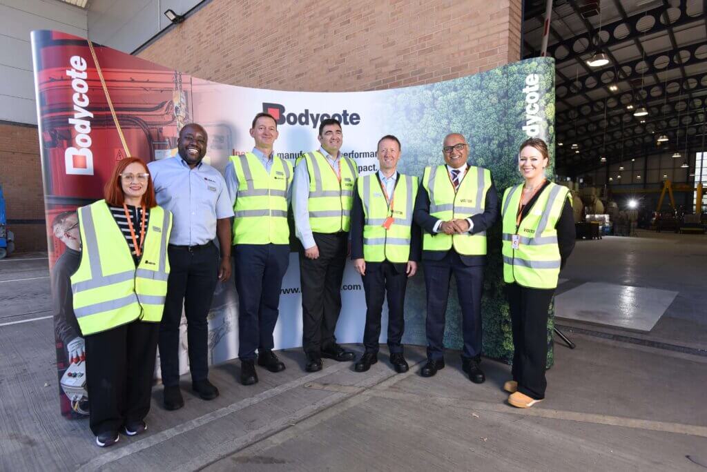 Seven people in yellow safety vests stand in front of a Bodycote display inside an industrial facility, smiling at the camera.