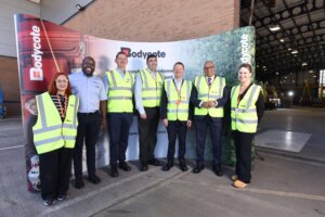 Seven people in yellow safety vests stand in front of a Bodycote display inside an industrial facility, smiling at the camera.