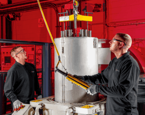 Two men wearing safety glasses and gloves operate industrial equipment at Bodycote, using a long tool to handle a metal cylinder during thermal processing in a red-lit facility.