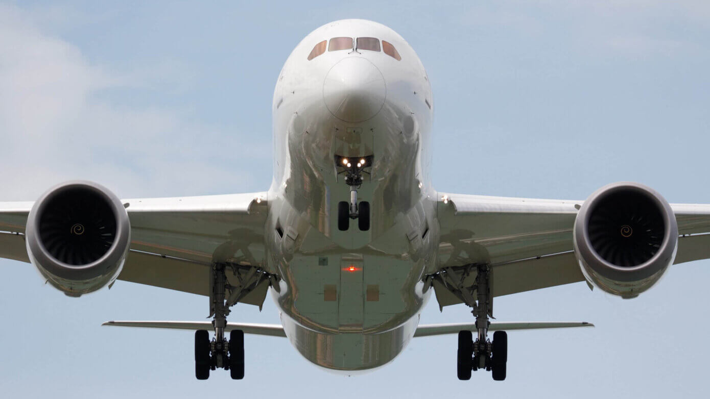 Forfra-billede af et stort passagerfly, der nærmer sig landingen med landingsstellet ude og begge motorer synlige mod en klar himmel.